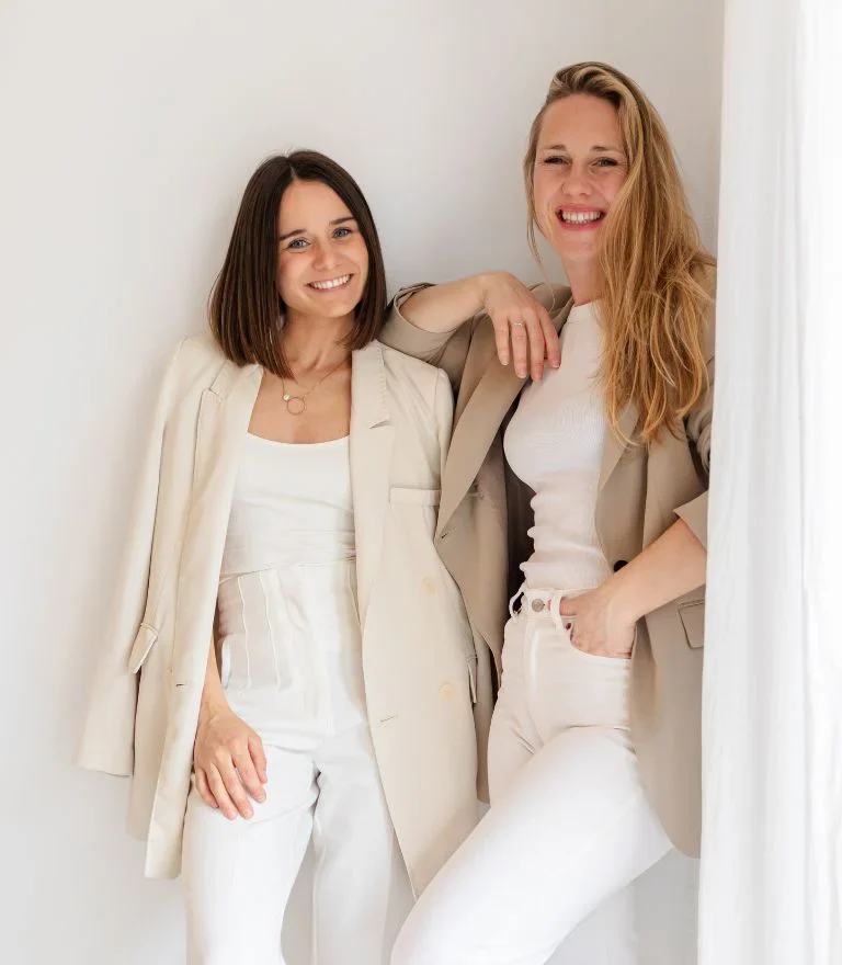 Zwei Frauen des Branddesign-Teams von Save Creations stehen gemeinsam vor einer weißen Wand in heller Kleidung.